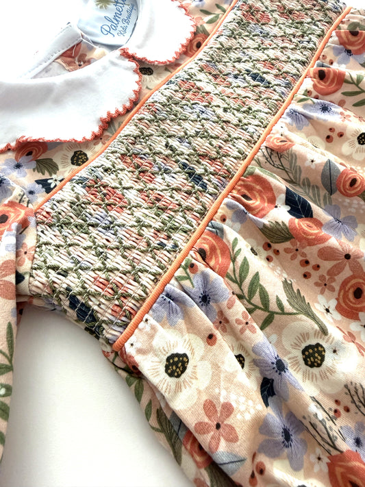 Close-up of a baby dress featuring a smocked bodice with peach piping, a white ruffled collar, and a floral skirt in peach, blue, and green on a cream background.