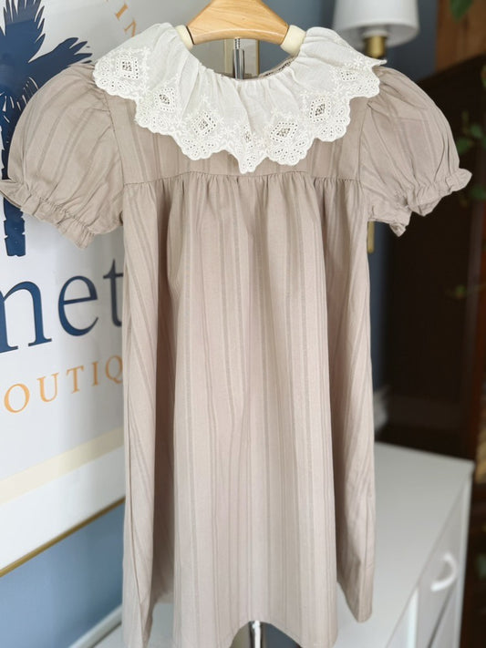 A beige short-sleeve baby dress with vertical pleats and a contrasting white lace ruffled collar, hanging on a wooden hanger.