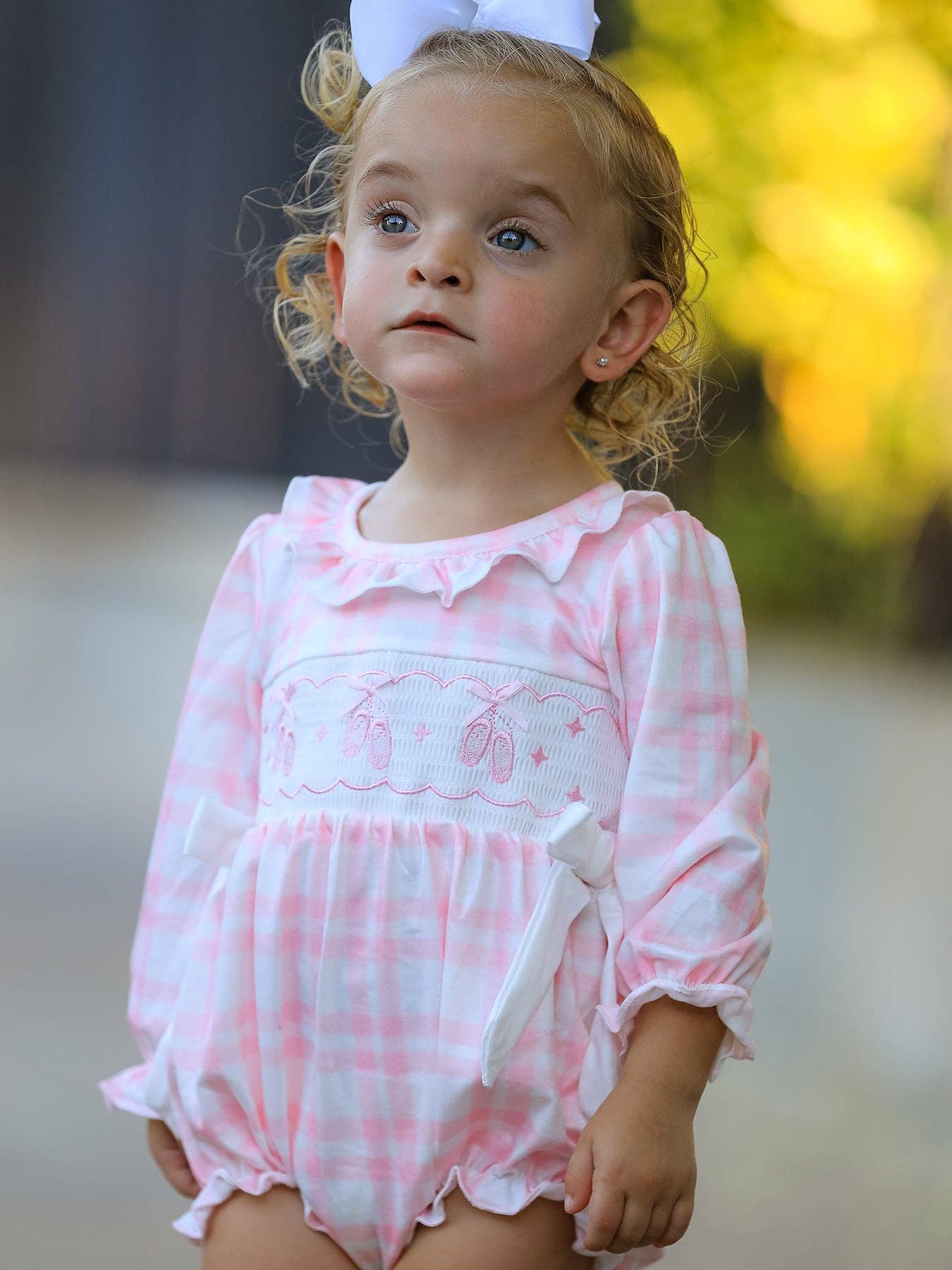 A toddler girl wearing a pink and white gingham long-sleeve romper with a smocked chest panel, ruffled collar, and white bow headband, photographed outdoors with a blurred background.