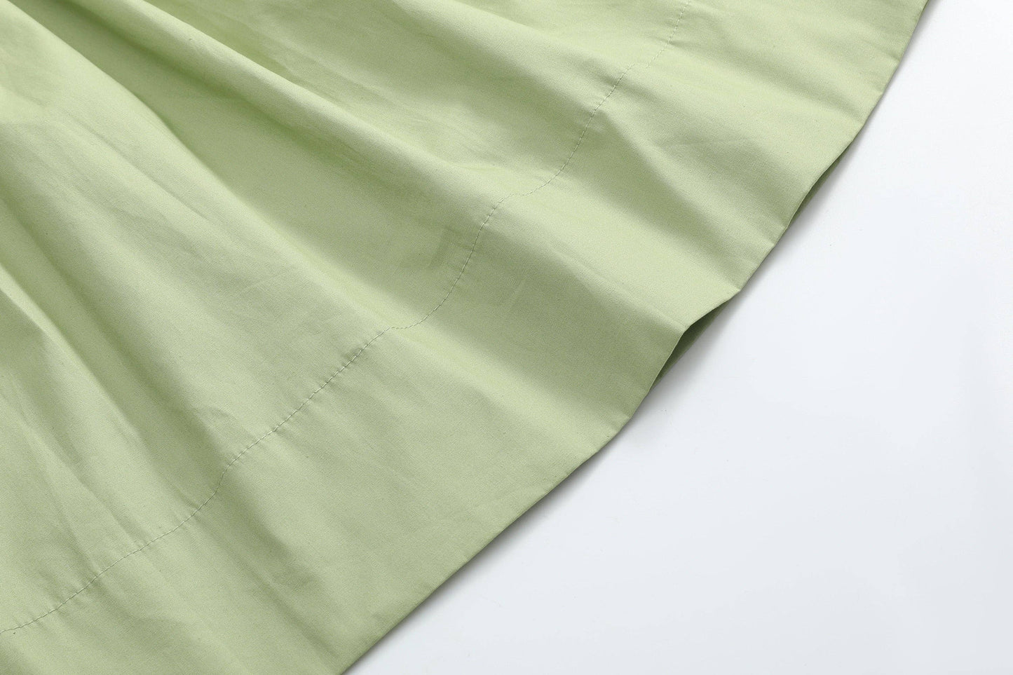 A close-up of pale sage green fabric with a visible fold and stitched edge, placed diagonally on a plain white background.