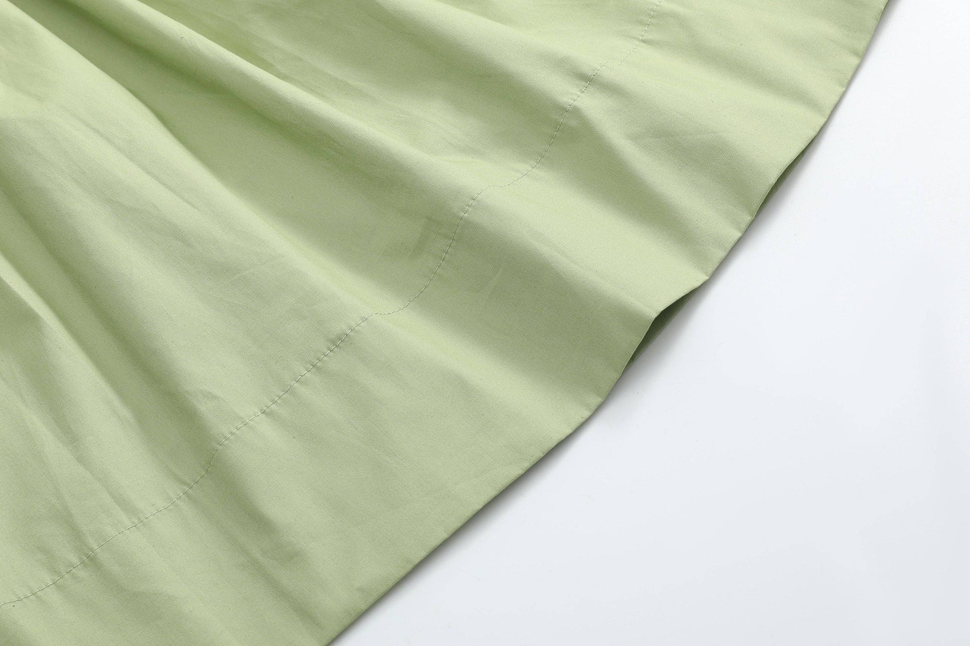 A close-up of pale sage green fabric with a visible fold and stitched edge, placed diagonally on a plain white background.