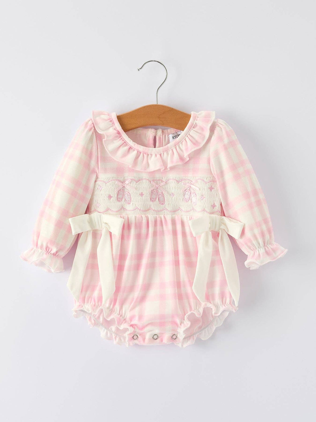 A pink and white gingham long-sleeve baby romper with a ruffled collar, embroidered chest detail, and cream ribbon bows, hanging on a wooden hanger against a light gray background.