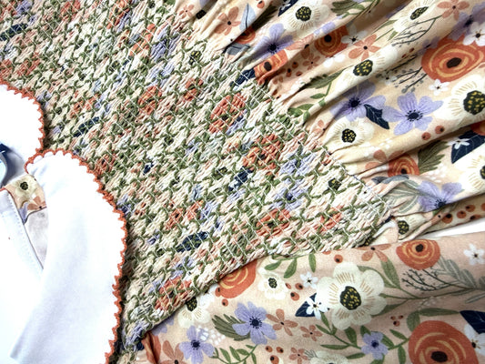 A close-up of a handmade quilted textile featuring a smocked floral pattern in green, orange, purple, and beige, layered with a solid cream fabric with orange scalloped edges.