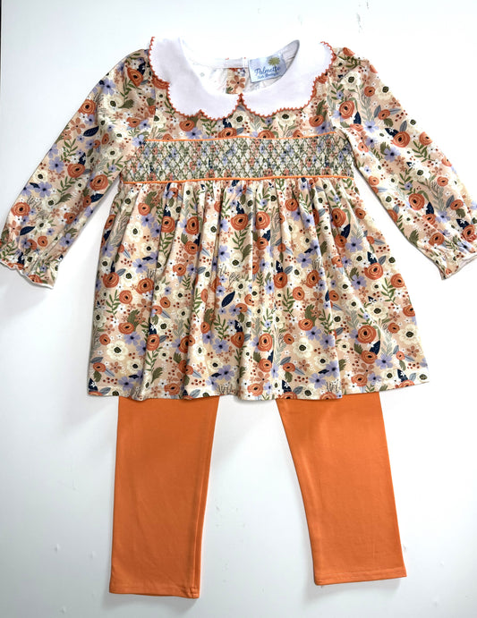 A toddler girl's two-piece outfit featuring a long-sleeve floral smocked dress with a white Peter Pan collar and orange-brown leggings.