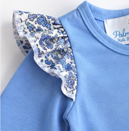 Close-up of a light blue children's top with a ruffled floral yoke in blue and white on the shoulder, showing part of the collar and a white neck label.