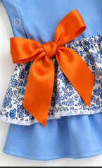 A close-up of a children's outfit featuring a light blue top with a large bright orange satin bow and a ruffled blue-and-white floral skirt.