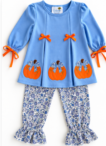 A toddler girl's two-piece outfit featuring a light blue long-sleeve top with orange pumpkin appliques and orange ribbon bows, paired with blue and white floral print ruffled pants.