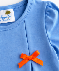 A close-up of a light blue baby top featuring a small orange bow on the chest and a visible 'Mellette de Barcelone' brand label inside the collar.
