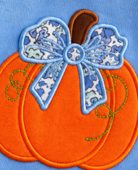 An embroidered patch of a pumpkin in bright orange with a large blue and white patterned bow on top and a brown stem, shown on a blue fabric background.