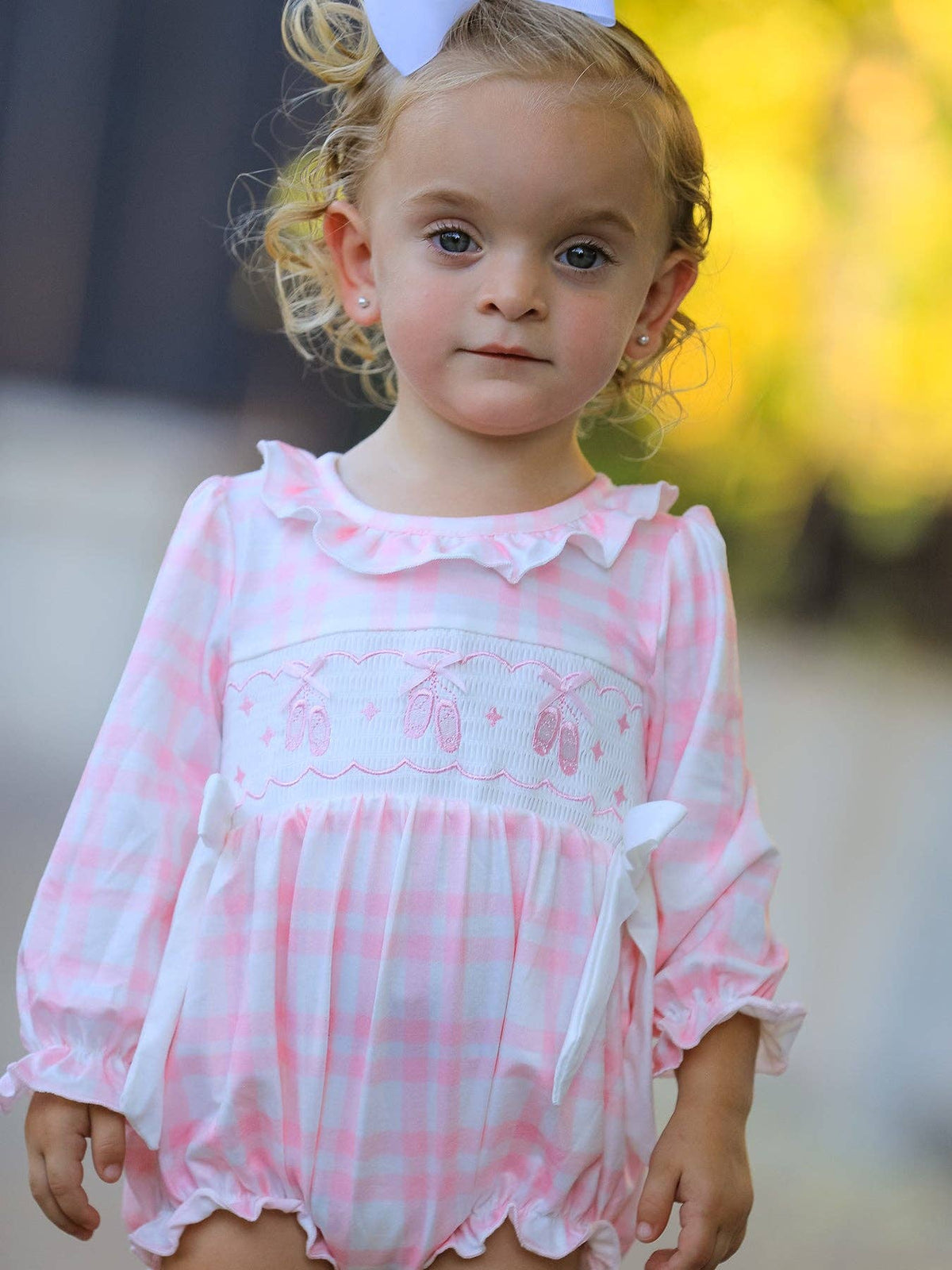 A toddler girl wearing a pink and white gingham long-sleeve romper with a ruffled collar, embroidered ballet dancers motif across the chest, and white bow details, photographed outdoors with a blurred yellow background.