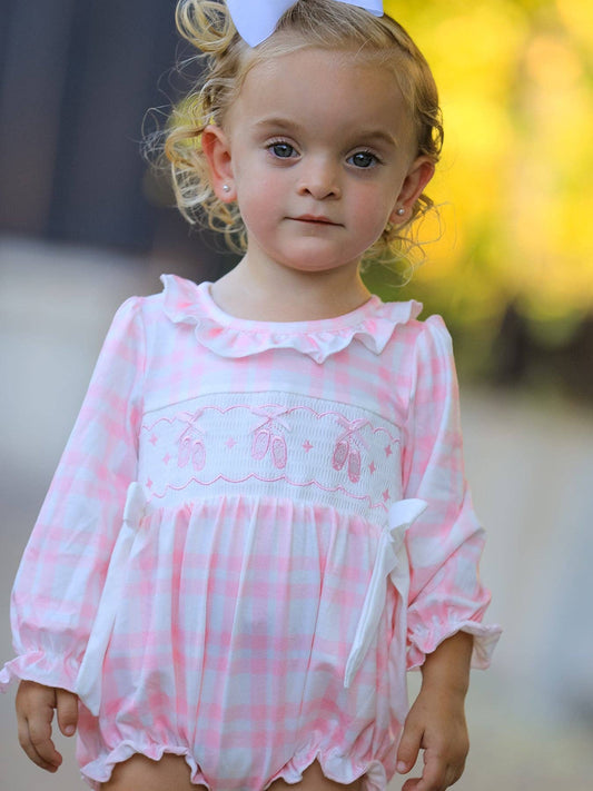 A toddler girl wearing a pink and white gingham long-sleeve romper with a ruffled collar, embroidered ballet dancers motif across the chest, and white bow details, photographed outdoors with a blurred yellow background.