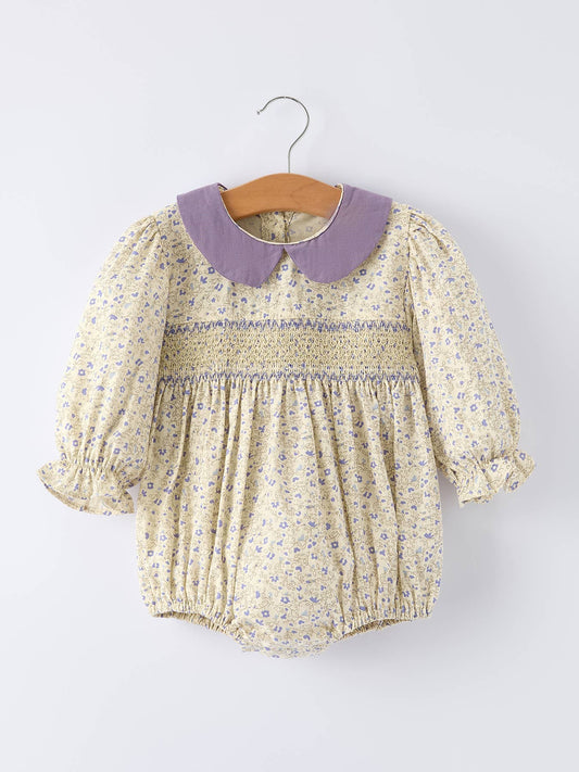 A baby girl's long-sleeve smocked romper in a cream floral print with a lavender purple Peter Pan collar, hanging on a wooden hanger against a light gray background.
