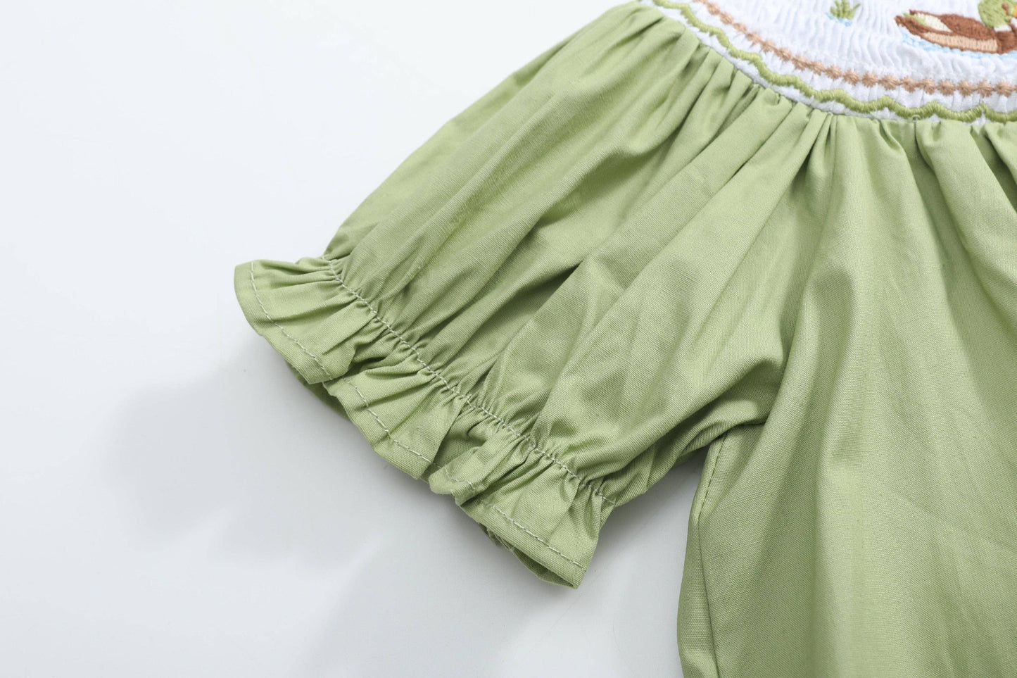 Close-up of a light olive green baby romper or dress with gathered ruffled sleeves and a white smocked bodice featuring embroidered details.